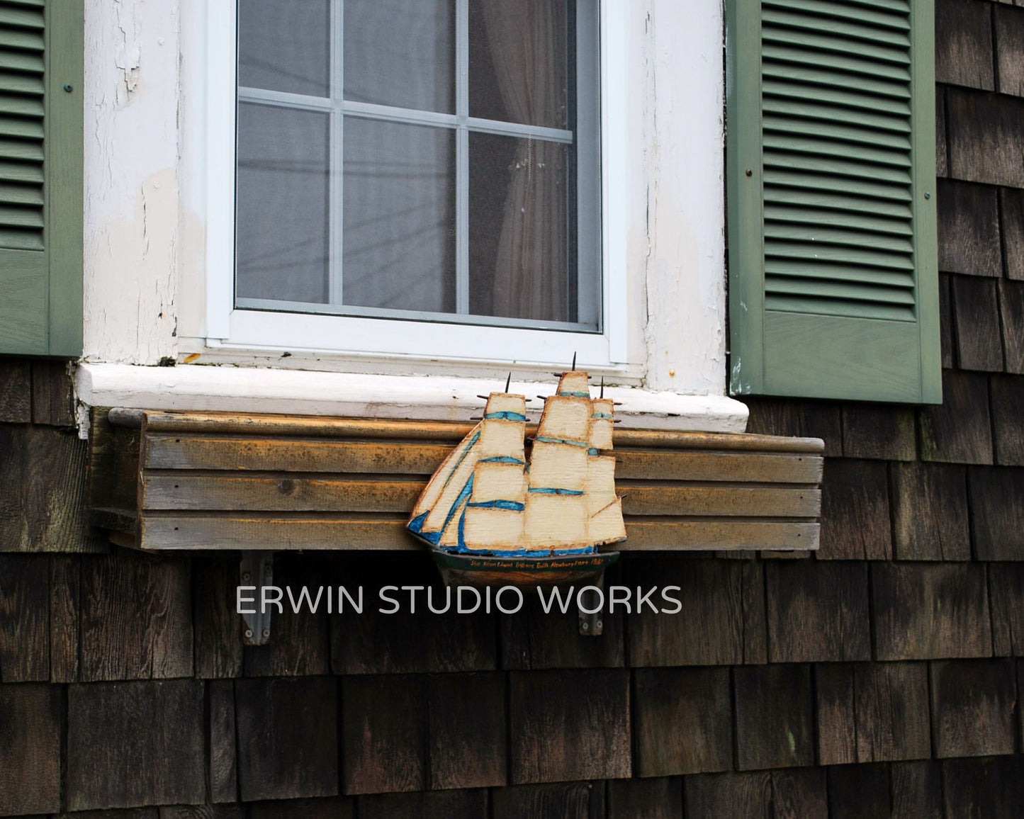 Wooden ship model on a window sill with green shutters and wooden wall.