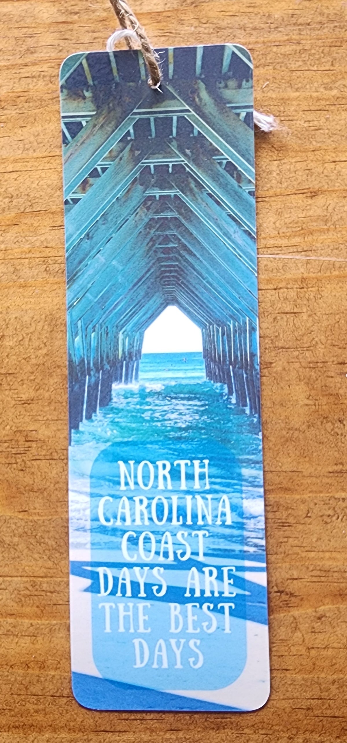 A rectangular paper bookmark or tag with a blue architectural tunnel photo and white text reading 'NORTH CAROLINA COAST ARE THE BEST DATES,' placed on a wooden surface.