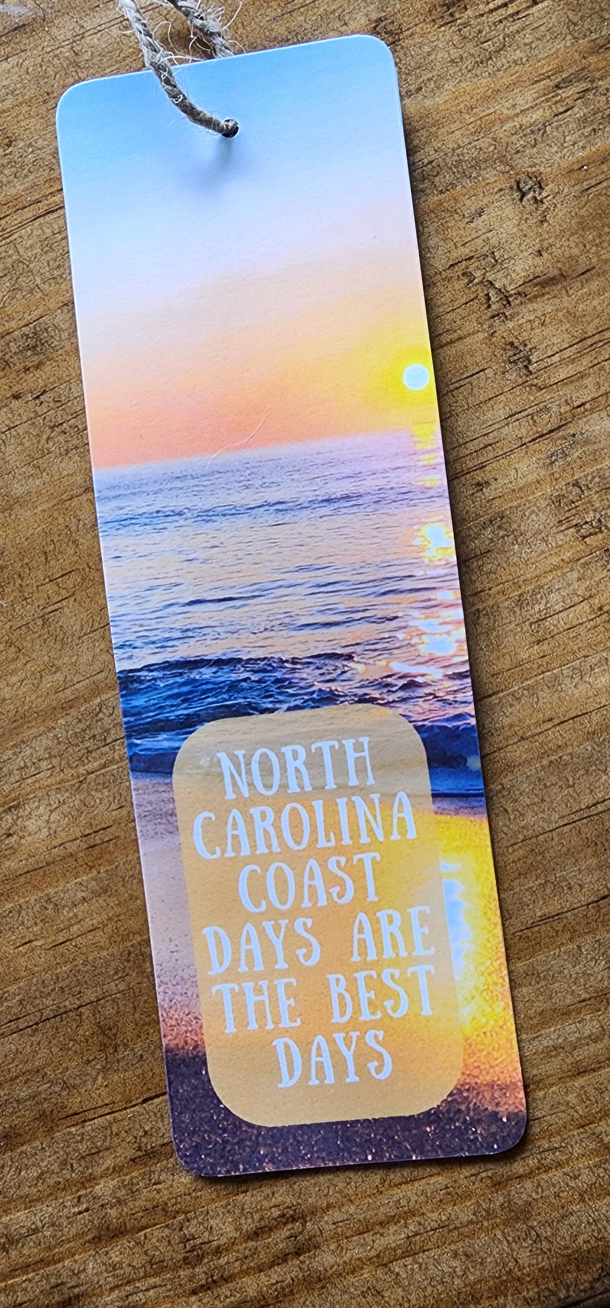 A rectangular printed bookmark or tag with a string hole, featuring a sunset coastal scene and the text 'NORTH CAROLINA COAST DAYS ARE THE BEST DAYS,' placed on a wooden surface.