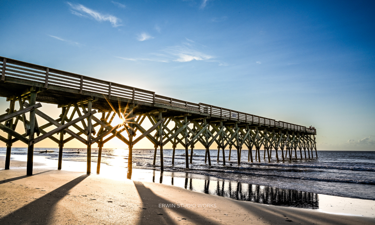 Wooden pier extending into the ocean with a clear blue sky and sun setting behind it. Wrightsville Beach, NC
