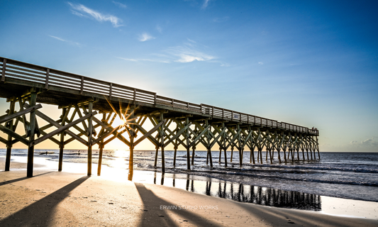 Wooden pier extending into the ocean with a clear blue sky and sun setting behind it. Wrightsville Beach, NC